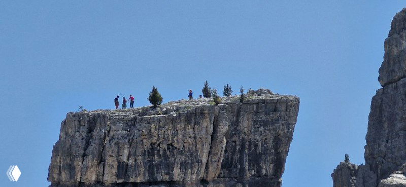 Небольшая плоская скала-утёс с людьми на краю; ощущение масштаба и открытого пространства в высокогорье Доломитов.