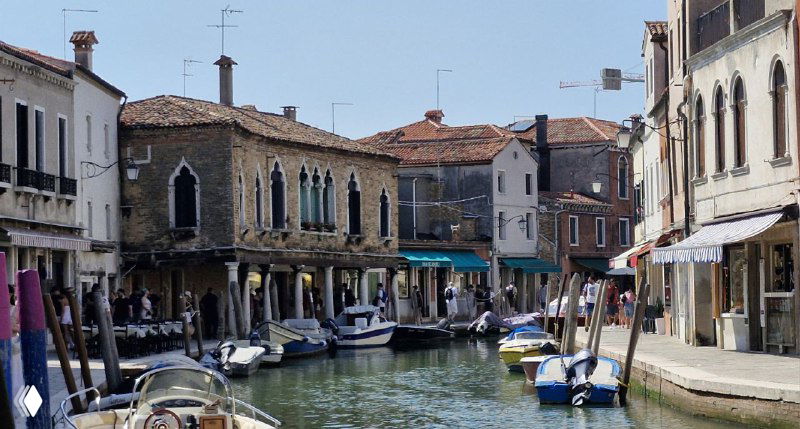 Узкий канал Мурано между старинными домами и магазинами, лодки пришвартованы у мостиков, тихая атмосфера острова.