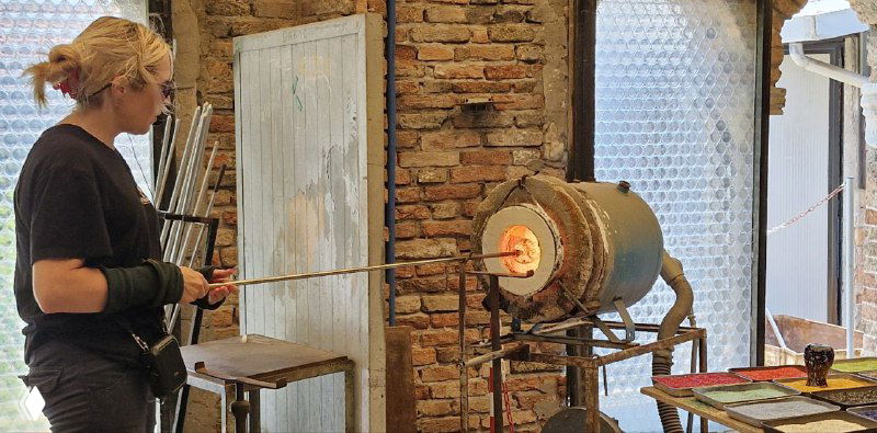 Женщина у печи формирует стеклянный предмет: рабочая сцена мастерской с инструментами и нагретой стеклянной массой на трубке.