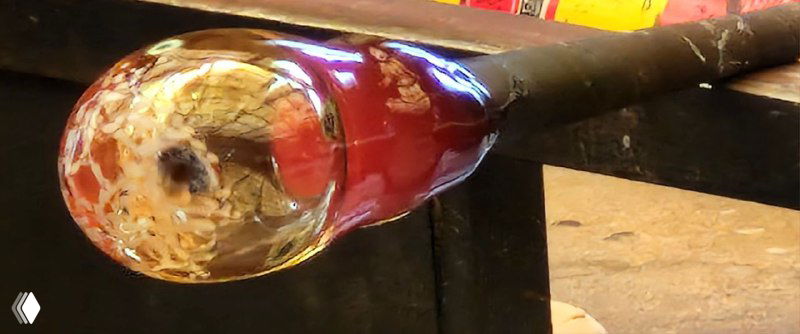 Конец стеклянной заготовки крупным планом: расплавленное цветное стекло на трубке перед формовкой и охлаждением, видна текстура и блеск.