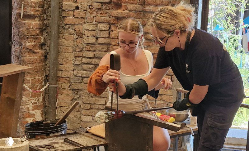 Две мастерицы работают совместно над формой: одна держит трубку, другая корректирует изделие холодными щипцами в мастерской.