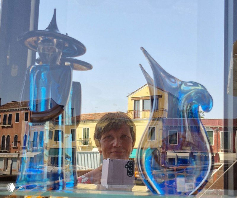 Витрина с муранскими изделиями: синие и прозрачные декоративные фигурки стоят на подоконнике и отражают фасады зданий за окном.