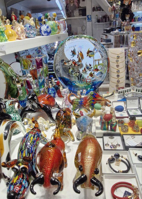Полка с множеством муранских сувениров и посуды: разноцветные стеклянные рыбки, вазы и декоративные тарелки в сувенирном магазине.