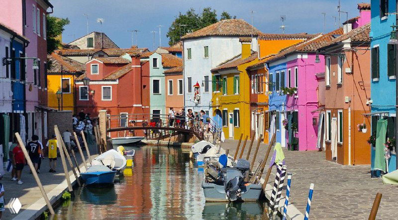 Узкий канал Бурано между рядами разноцветных домов, небольшие лодки у причалов, пешеходные дорожки и типичная атмосфера островного поселения.