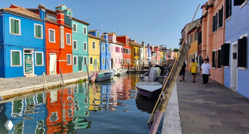 Пешеходная набережная Бурано с рядами ярко окрашенных домов, туристы и местные жители проходят мимо маленьких причалов и лодок у воды.