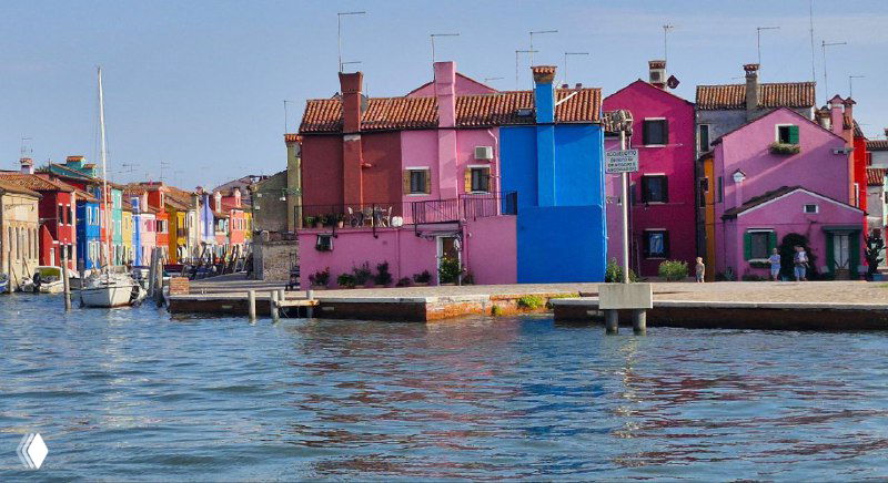 Вид с воды на буранские фасады: розовые и синие дома у причала, лодки и морская атмосфера, характерный портовый пейзаж маленького острова.