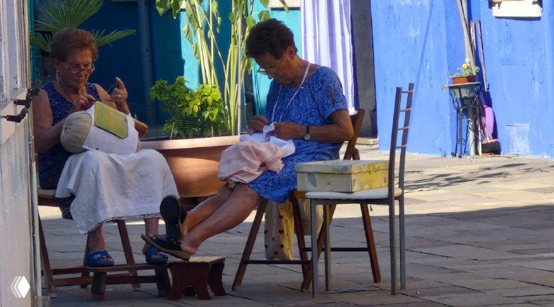 Две буранские кружевницы за работой на улице: пожилые женщины плетут кружево вручную, традиционная техника и сцена ремесленного мастерства на острове.