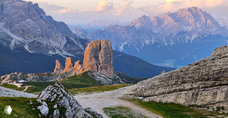 Скалистая формация и дорога на переднем плане в закатном свете, с видом на удалённые хребты и мягкую альпийскую растительность.