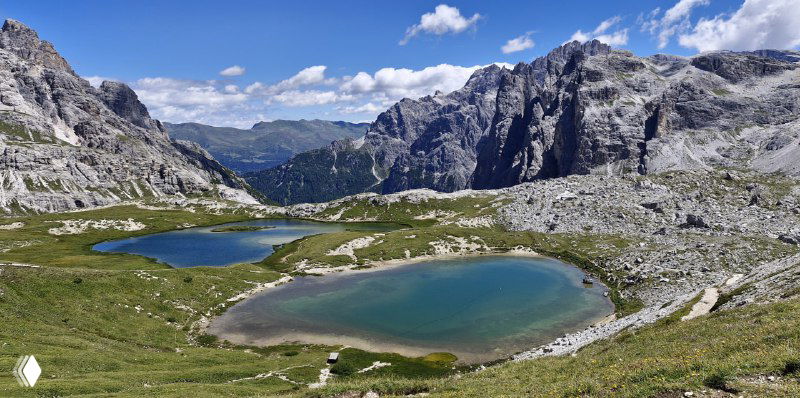 Две бирюзовые горные лагуны в каменистом цирке, вокруг видны тропы и седловины — типичный альпийский озёрный пейзаж Доломит.