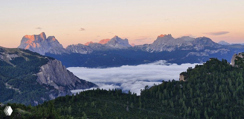 Широкая панорама с морем облаков в долине и силуэтами дальних вершин, тёплые оттенки заката на горном хребте.