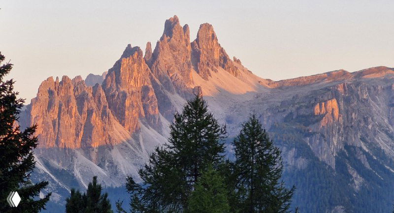 Залитые тёплым светом пики Доломитов на фоне лиственниц внизу; ощущение глубины и вечерней подсветки горного массива.