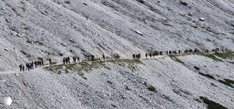 Длинный горный троп с группами туристов, выстроившимися по каменистой поверхности; показывает людность популярных маршрутов и троп.