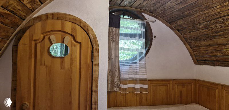 Интерьер хоббитского домика: деревянная дверь с круглым иллюминатором, простая отделка и окно с занавеской — спартанская уютность.