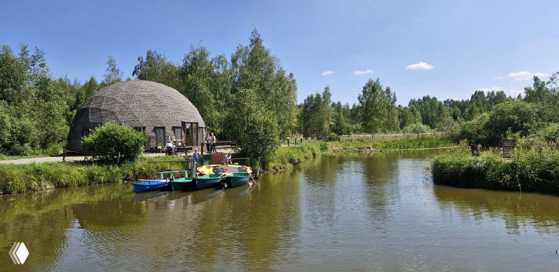 Озеро на территории Хоббитлэнда в Подмосковье: плоскодонные лодочки у берега, купольные строения и зелёная прибрежная растительность.
