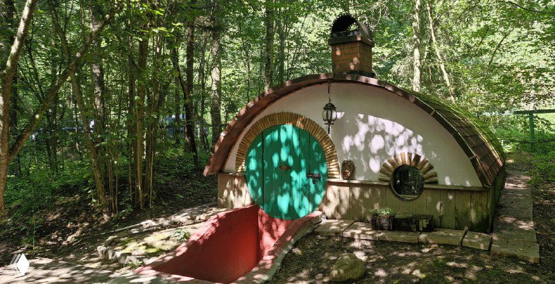 Маленький хоббитский домик с круглой зелёной дверью и красной подпоркой у входа, утопающий в тенистом лесном окружении Подмосковья.
