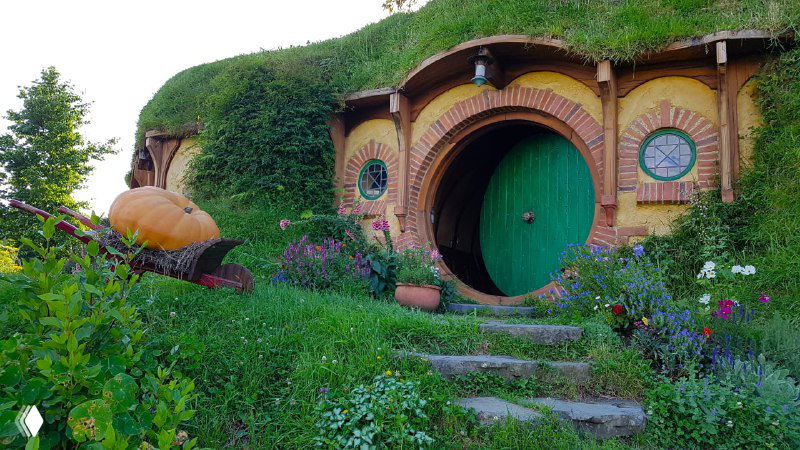 Хоббитон в Новой Зеландии: домик с зелёной круглой дверью, цветами и аккуратно выложенной лестницей, типичный пейзаж киностудийного комплекса.