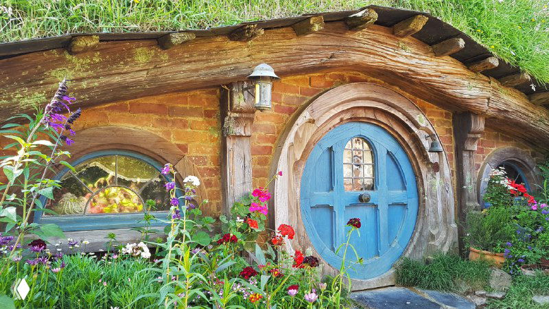 Крупный план фасада хоббитского домика в Новой Зеландии с голубой круглой дверью и яркими цветами у входа, декоративная атмосфера.