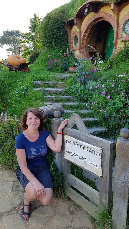 Автор на фоне хоббитской входной зоны в Хоббитоне (Новая Зеландия), сидит у таблички на фоне цветущего уголка киностудии и домиков.