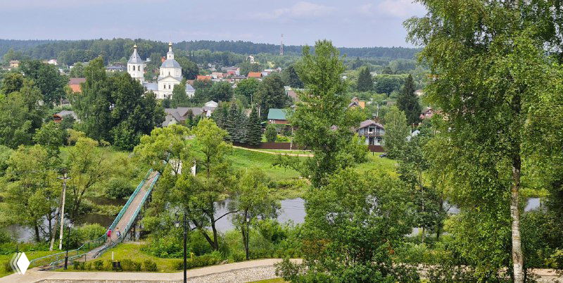Панорамный вид с высоты на речку, детскую горку и силуэты церкви вдалеке; типичный сельский пейзаж Подмосковья с зеленью и домами.