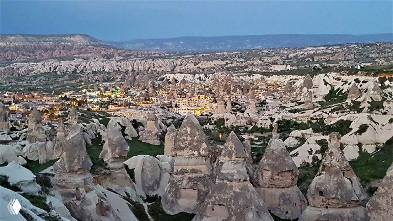 Рассветный или сумеречный вид на плотную группу перибаджалары и туфовых образований с городскими огнями Гёреме вдалеке, подчёркивающий масштаб местности.