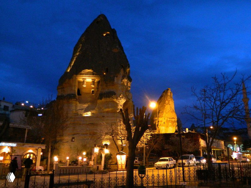 Крупный ночной снимок одной из вырубленных в туфе башен или домов Гёреме с тёплым светом из окон и уличными элементами на переднем плане.
