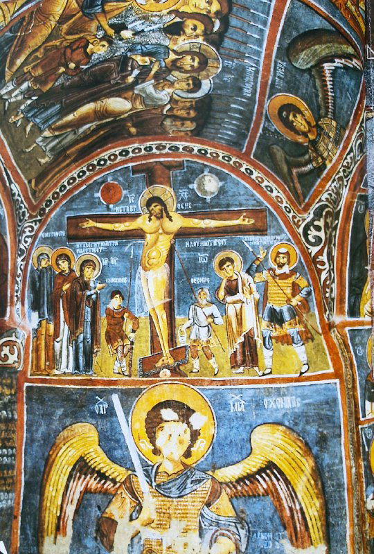 Вертикальная фреска с распятием и фигурами святых в пещерной церкви; охристые и синие тона, архитектурные мотивы и рельефность изображения.