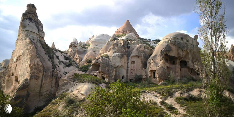 Группа высеченных в скале жилищ и пещер на холме у подножья, видны окна и ниши в массиве, растительность по склонам.