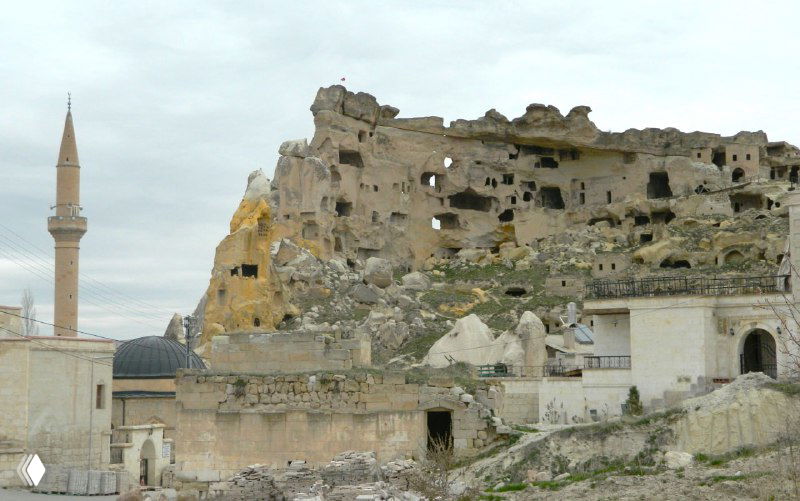 Каменные древние постройки и обширный скальный массив у подножья поселка, на переднем плане минарет и жилые здания на фоне сводов.