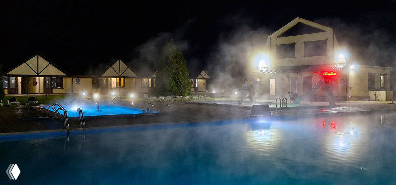 Вечерние термальные бассейны с паром и огнями на территории комплекса; коттеджные корпуса и дорожки вокруг воды создают уютную атмосферу.