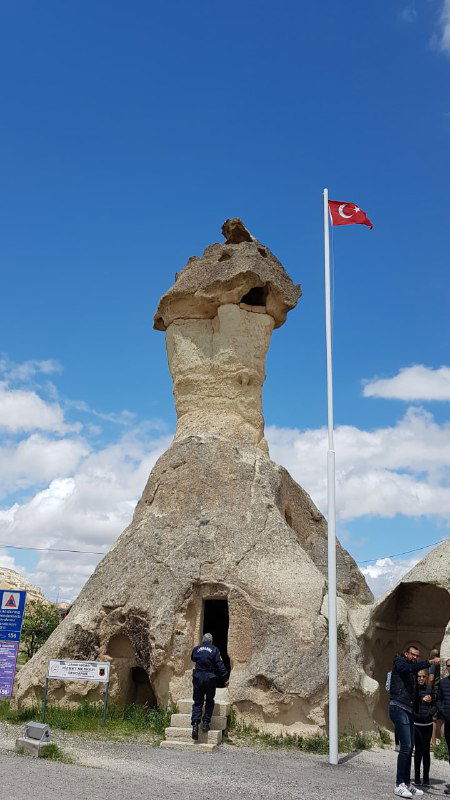 Каменное строение с выдолбленным входом и флагом Турции на ходу; на переднем плане люди, подходящие к достопримечательности.
