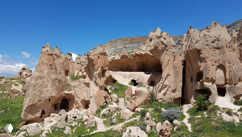 Панорамный вид на разрушенные и частично сохранившиеся жилищные скалы Зельве, уступы и зелёные участки внизу на фоне голубого неба.