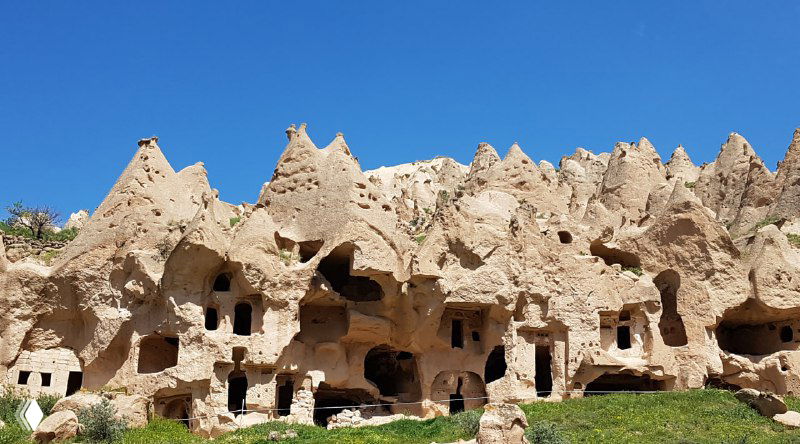 Остроконечные скальные шпили и фасады с окнами-отверстиями, ряд конических форм Каппадокии на переднем плане под ясным небом.