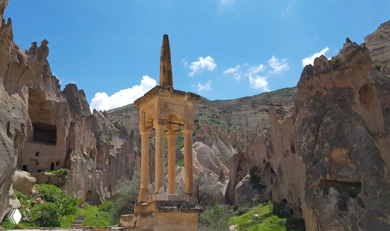 Каменная колонна или руинированный памятник среди скал Зельве: архаичный архитектурный элемент в окружении пещерных выемок и зеленых склонов.