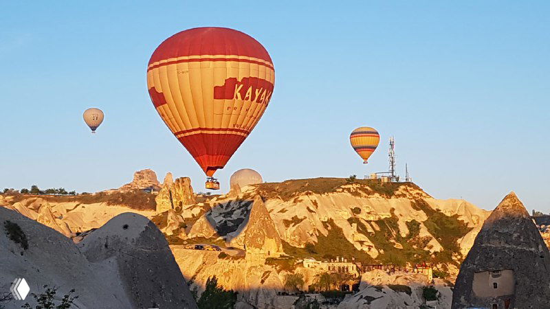 Крупный цветной воздушный шар в полёте над скалистым ландшафтом Каппадокии, свет рассвета подчёркивает текстуру вулканических пород.