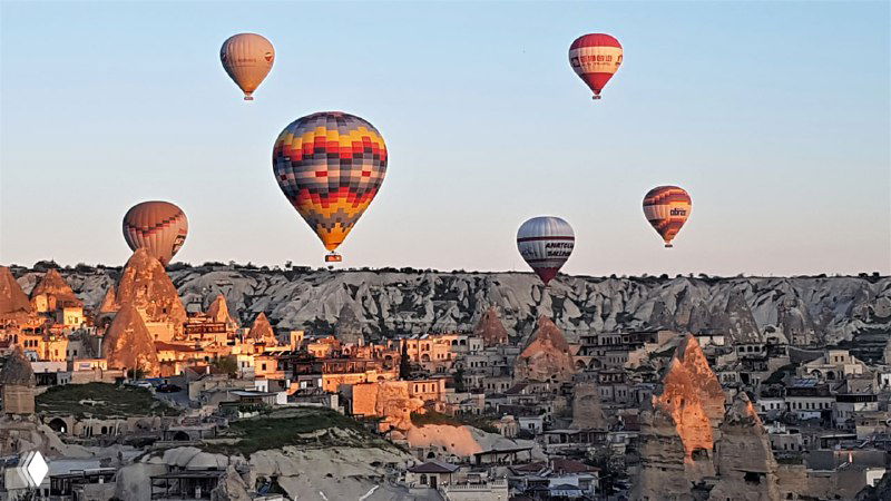 Горизонтальный кадр: несколько воздушных шаров над панорамой Гёреме и характерными «феерическими трубами» и домами в скалах.