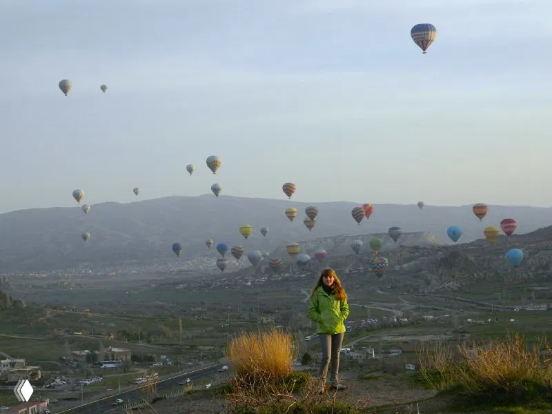 Широкий кадр с большого расстояния: множество воздушных шаров над расчерченным рельефом Каппадокии на фоне гор и горизонта.