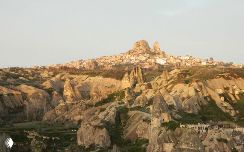 Тёплый вечерный свет освещает туфовые формации Каппадокии и силуэт крепости Учхисар на вершине холма с удалённым пейзажем.