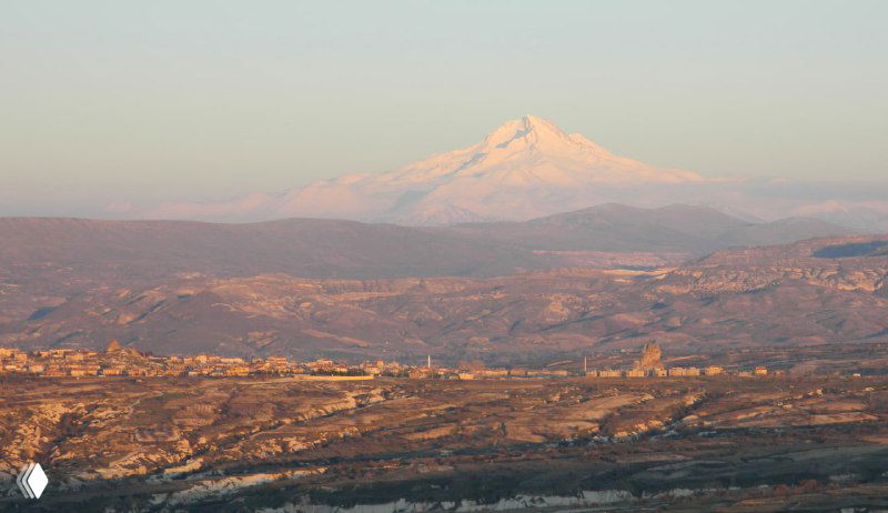 Вдали виден заснеженный конус вулкана Эрджияс/Хасан на фоне равнины — вид, который открывается из Учхисара в ясную погоду.
