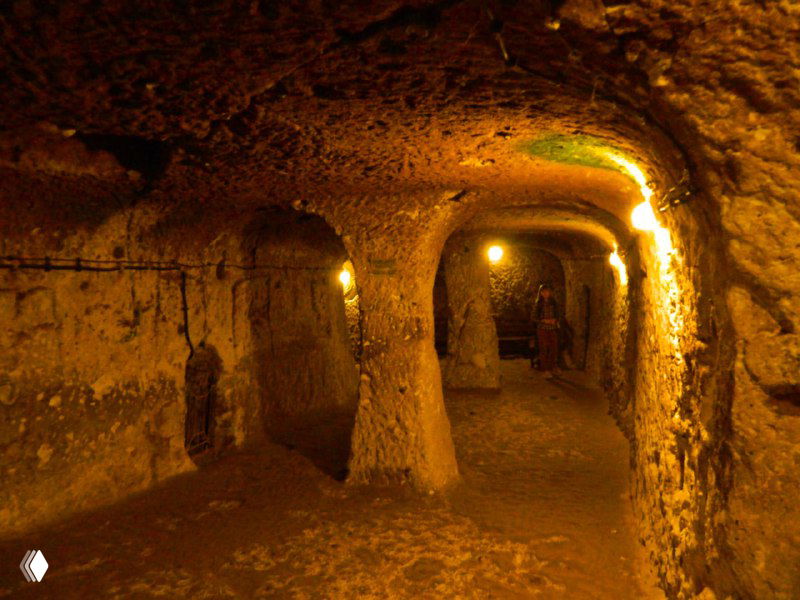 Освещённый коридор Деринкую с колонной-подпорой посередине, вырубленные дверные проёмы и ровный пол — вид туристического маршрута внутри города.