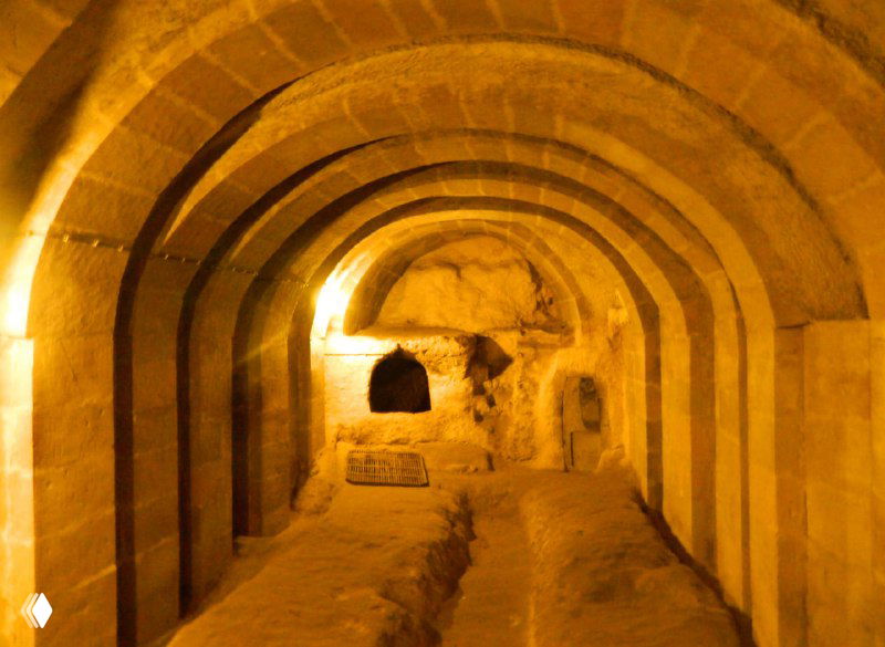 Арочный проход внутри Деринкую с вырезанными нишами и ровным полом, тёплое освещение подчёркивает структуру помещений и архитектуру выемок.