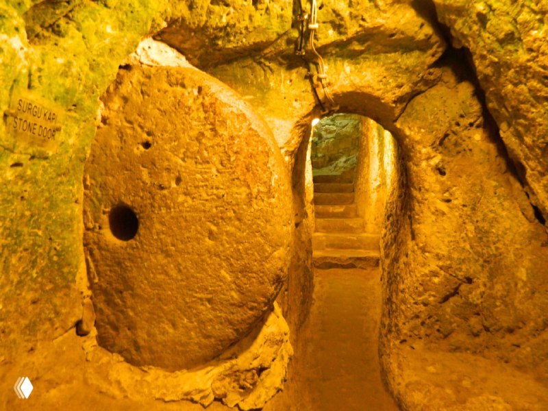 Каменное колесо-дверь в узком тоннеле Деринкую: массивный круг с отверстием в центре, готовый выкатываться и перекрывать вход в город.