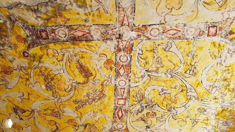Крупный план потолочной фрески с крестом и растительными орнаментами в жёлто-красной гамме внутри церкви Кешлик, заметны следы копоти и времени.