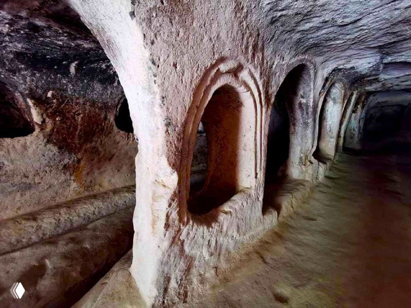 Длинный внутренний коридор монастыря Кешлик с арочными нишами и выемками в туфе, видны жилые и хозяйственные помещения, не подготовленные для массового туризма.