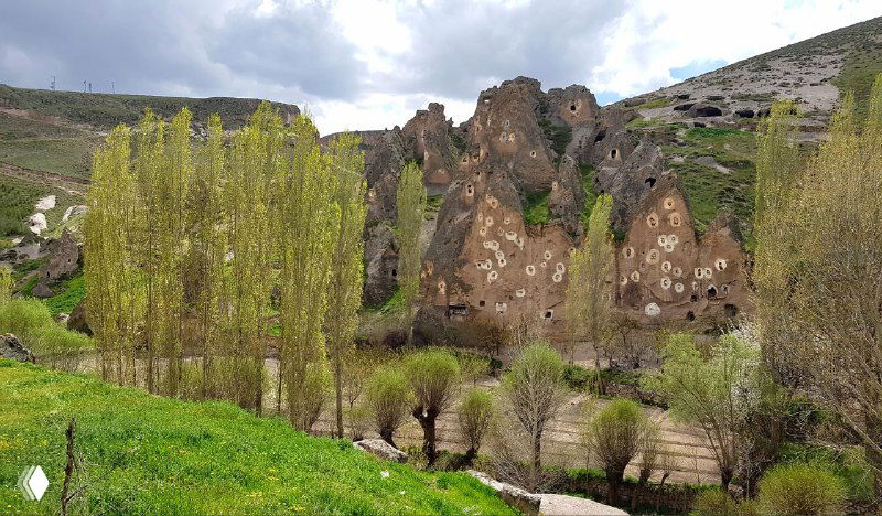 Ущелье Соганлы с туфовыми скалами и множеством белых отметок вокруг входов в голубятни, весенний зелёный ландшафт.