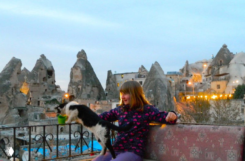 Девочка на балконе пещерного отеля в Каппадокии играет с кошкой на фоне туфовых формирований и огней поселка.
