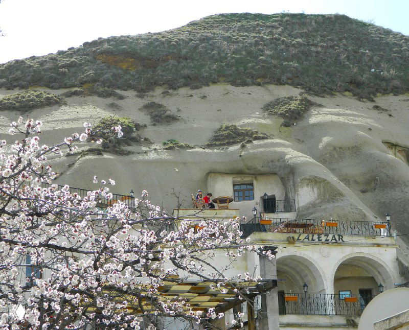 Фасады старинных построек и пещерных отелей на склоне, цветущие деревья на переднем плане и входы в небольшие термальные лавки.