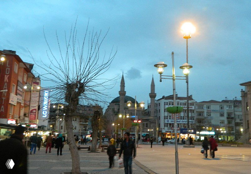 Площадь в вечернем освещении с уличными фонарями, людьми и силуэтом минарета — кадр ожидания автобуса в городском центре.