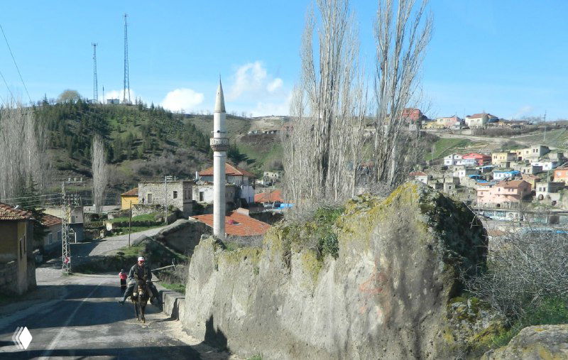 Деревенский пейзаж с каменной скалой, минаретом и домами на склоне; вид из окна автобуса во время переезда по региону.