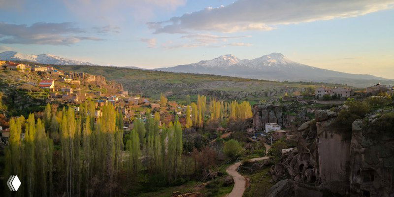 Долина с деревьями и домами, тропа вдоль окраины поселка, на заднем плане плато и заснеженные вершины — широкий план ландшафта региона.