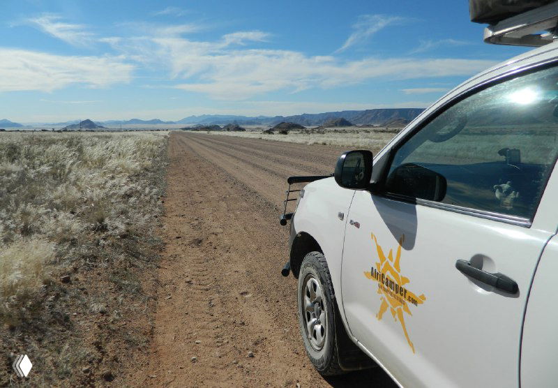 Внедорожник на просёлочной дороге пустынной местности: вид сбоку автомобиля с палаткой на крыше, ровный грунтовый путь и низкая кустарниковая растительность вокруг.